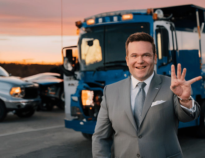 A politician, in a tailored suit, gestures proudly beside a gleaming tow truck at dusk.